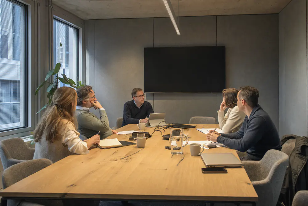 Vergadering aan een eiken tafel in een ruimte met grijze viltpanelen op de wanden